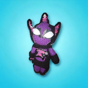 May include: Purple and black crocheted plush toy with a white horn and a black mask. The toy has a pink and black design on its chest.