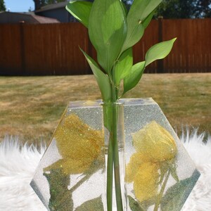 May include: A clear hexagonal vase with gold glitter embedded in the resin. The vase has a single opening for flowers and is filled with yellow glitter roses. A green plant is in the vase.