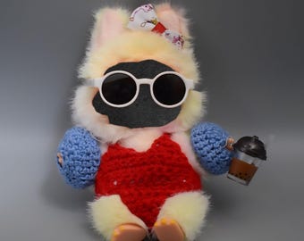 May include: A plush toy with a black face, wearing white sunglasses, a red crocheted swimsuit, and blue crocheted mittens. The toy has a yellow and pink fur body and a patterned bow. It holds a small boba tea.