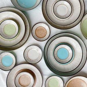 Puede incluir: Vista aérea de una colección de platos y cuencos de cerámica. Los platos tienen una base de color crema con finas líneas marrones verticales. Algunos cuencos tienen un centro de color sólido en tonos de azul, verde y marrón. Los platos varían en tamaño.