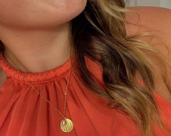 May include: Gold-toned necklace with a circular pendant and a small pearl drop. The pendant has a textured design. The necklace is worn over an orange halter top.