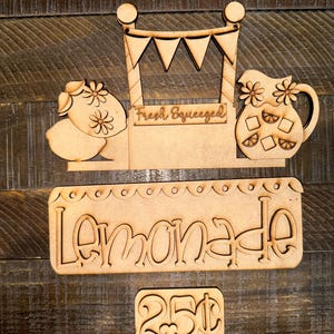May include: Wooden lemonade stand kit with cut-out shapes. Includes a sign that says "Lemonade", a sign that says "Fresh Squeezed", a pitcher, lemons, and a price tag that says "25¢".