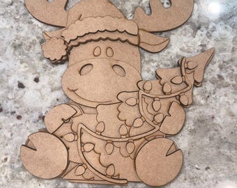 Puede incluir: Decoración navideña de madera con un alce con gorro de Papá Noel y un árbol de Navidad con luces. El alce está sentado y tiene una ranura para un soporte. La decoración es de madera sin terminar.