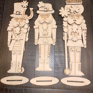 May include: Three wooden nutcracker ornaments with a St. Patrick's Day theme. Each nutcracker is painted in shades of brown and features a green shamrock, a green hat, and a white beard. The nutcracker on the left is holding a horseshoe, the middle nutcracker is holding a shamrock, and the nutcracker on the right is holding a pot of gold.