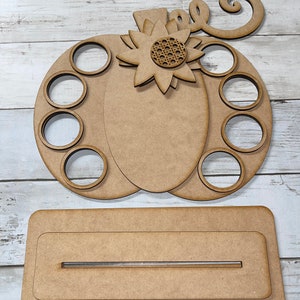 May include: Unpainted wooden pumpkin-shaped craft with eight round cutouts and a sunflower design. The pumpkin is attached to a separate wooden base with a slot for a dowel.