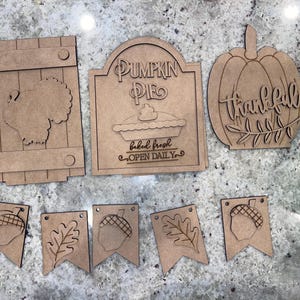 May include: A set of wooden craft signs with a Thanksgiving theme. The signs feature a turkey, a pumpkin pie, a pumpkin with the word "thankful", and a banner with acorns and leaves.