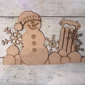 May include: Wooden cutout of a snowman with a hat and scarf, standing on a base with snowballs and a sled. The snowman has a carrot nose and three buttons. There are snowflakes around the snowman.