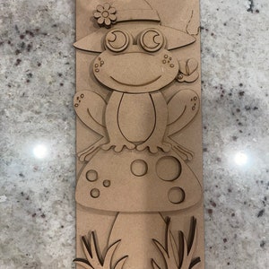May include: Wooden cutout of a frog wearing a hat and sitting on a mushroom. The frog has large eyes and a smile. The mushroom has white spots. The cutout is on a brown background.