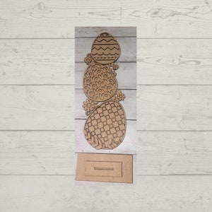 May include: A wooden Easter egg stand with three stacked eggs. The eggs are decorated with different patterns, including flowers, geometric shapes, and bunny silhouettes. The stand has a slot for inserting a card or message.