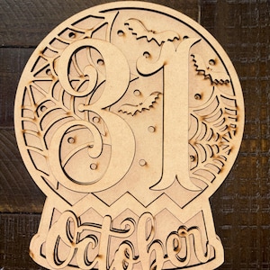 May include: A laser-cut wooden sign shaped like a snow globe featuring the number 31 in large script, the word October in cursive script at the base, and decorative cutouts of spider webs and flying bats throughout the circular design.