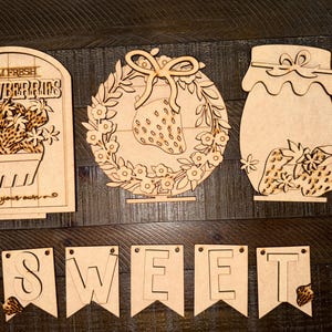 May include: Wooden craft kit for a strawberry themed sign. The kit includes a sign that says "Farm Fresh Strawberries, pick your own", a wreath with a strawberry in the center, a jar with strawberries, and a banner that says "SWEET".