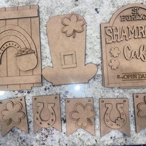 May include: A set of unfinished wooden cutouts for a St. Patrick's Day craft project. The set includes a leprechaun hat, a pot of gold, a rainbow, a shamrock, a horseshoe, and a sign that says "St. Patrick's Shamrock Cafe Open Daily".