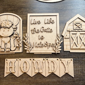 May include: A collection of unfinished wooden cutouts featuring a Highland cow, a barn, a sign with the text "Live life the Gate is Wide Open", and a banner spelling "HOWDY". These rustic decorations are perfect for DIY projects.