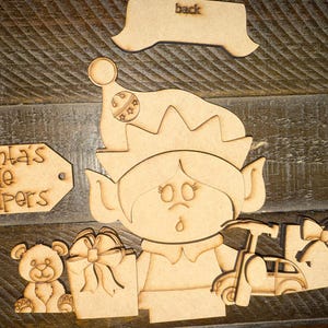 May include: Wooden cutouts of a Christmas elf, a teddy bear, gifts, and a car. The elf wears a hat with a bell and stars. The text "Santa's little helpers" is on a tag. The word "back" is on a separate cutout.