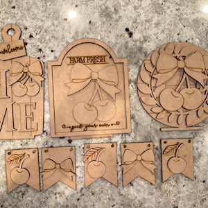 May include: A set of unfinished wooden cutouts featuring cherries and bows. The set includes a "Home" sign, a "Farm Fresh" sign, a wreath, and four banner flags. The signs and flags are ready to be painted or stained.