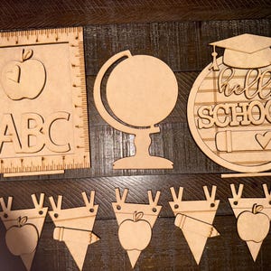 May include: A collection of unfinished wooden school-themed cutouts. Includes an apple and ABCs on a ruler, a globe, a circular sign with a graduation cap and the words "hello SCHOOL", and several pennant-shaped cutouts with school-related images.
