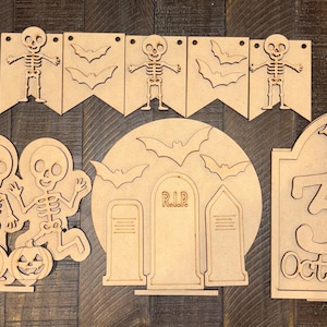 May include: A set of unfinished light brown wooden laser-cut decorations featuring skeletons, bats, pumpkins, tombstones, and a sign that reads 31 October with a spider web and spider. The pieces include a banner, standing figures, and plaques.