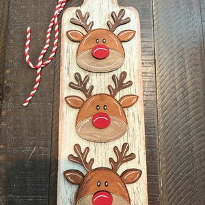 May include: A rectangular wooden wall hanging with a distressed white finish, featuring three layered reindeer heads with brown antlers, tan faces, and bright red noses. A red and white twisted rope is attached to the top for hanging purposes.