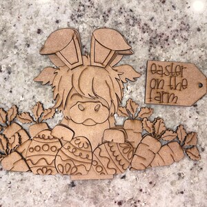 May include: Wooden cutout of a Highland cow with bunny ears holding Easter eggs. The cow is surrounded by carrots and greenery. A wooden tag reads "Easter on the farm".