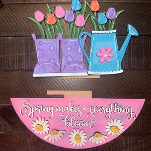 May include: A wooden spring-themed decoration featuring a pink semi-circle with the text "Spring makes everything bloom" and daisy accents. Above, are purple rain boots and a blue watering can, each holding colorful tulips.