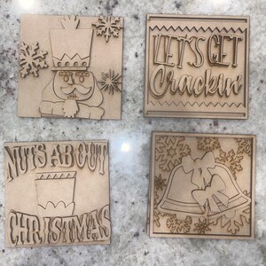 May include: Four wooden Christmas signs with different designs. The first sign features a nutcracker with a crown and a mustache. The second sign says "Let's Get Crackin". The third sign says "Nuts About Christmas". The fourth sign features two bells with bows and snowflakes.