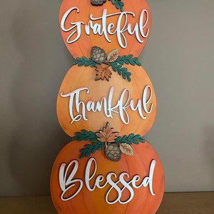 May include: A stack of three orange pumpkins with green leaves and pine cones. The pumpkins are painted with the words "Grateful", "Thankful", and "Blessed" in white lettering.