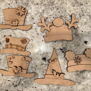 May include: A collection of unfinished wooden hat cutouts, including a leprechaun hat, a hat with antlers, a witch hat with bats, and a hat with a snowflake design. These decorative cutouts are ideal for crafting and seasonal decor.