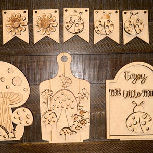 May include: Wooden cutouts of various shapes, including banners with flower and ladybug designs, a mushroom, a cutting board with mushroom and ladybug designs, and a sign that says "Enjoy the little things."