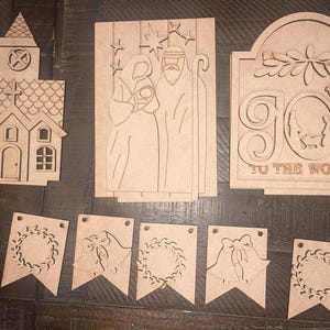 May include: A set of unfinished wooden Christmas decorations, including a church, a nativity scene, a "Joy to the World" sign, and five banner flags. The flags feature a wreath, bells, and a wreath.