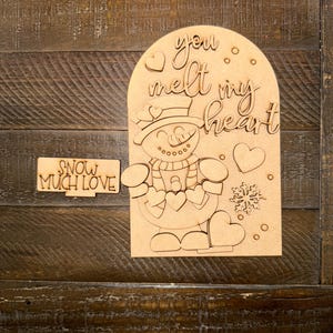 May include: A wooden snowman-themed decoration with the phrase "you melt my heart" and a smaller sign that says "snow much love". The snowman has a hat, scarf, and holds hearts. A snowflake and hearts are also included.