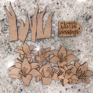 May include: Wooden Easter-themed cutouts, including lilies, grass, and a tag with the words "Easter blessings." The cutouts are made of light brown wood and are arranged on a speckled surface.