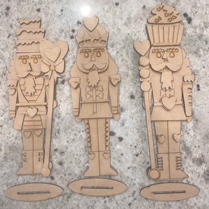 May include: Three unfinished wooden nutcracker ornaments with a Valentine's Day theme. The nutcrackers are decorated with hearts and have a whimsical design.
