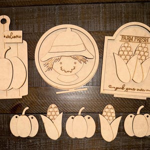 May include: A collection of unfinished wooden fall-themed cutouts. Includes a cutting board with a pumpkin, a scarecrow, a sign that says "Farm Fresh" with corn, and several pumpkins and corn cutouts. The word "welcome" is on the cutting board.