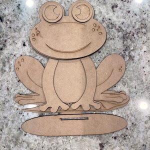 May include: A wooden frog cutout with large eyes and a wide smile. The frog is sitting on a wooden base.