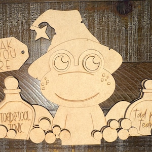 May include: Wooden cutout of a frog wearing a witch hat with a star. The frog is flanked by two potion bottles labeled "Toadstool Tonic" and "Tad Pole Tears". A tag reads "Croak If You Dare".
