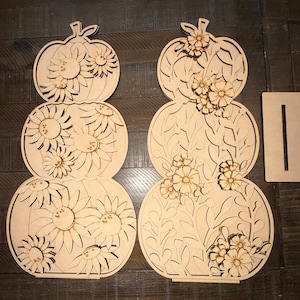 May include: Two laser-cut wooden pumpkin-shaped decorations featuring stacked segments. The left pumpkin is adorned with sunflower cutouts, while the right pumpkin features floral and vine patterns. A rectangular wooden base with a slot is included.