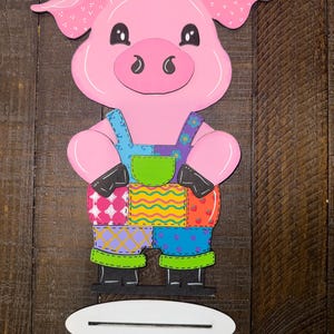 May include: A pink pig-shaped wooden cutout wearing patchwork overalls with blue straps and green trim. The pig has black eyes, ears with white polka dots, and a white base for standing. The pig is a decorative item.