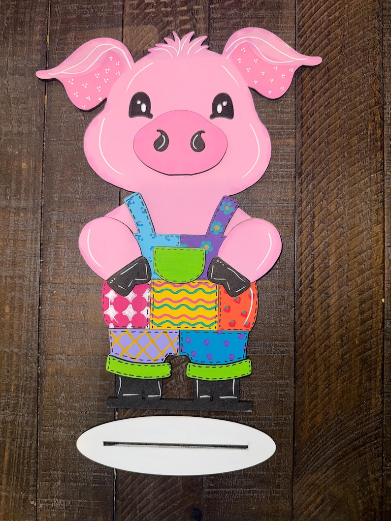 May include: A pink pig-shaped wooden cutout wearing patchwork overalls with blue straps and green trim. The pig has black eyes, ears with white polka dots, and a white base for standing. The pig is a decorative item.