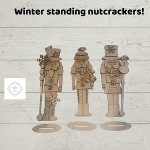 May include: Three wooden winter-themed standing nutcrackers with individual bases. Each nutcracker is a different design, with snowflake and winter hat details. The text "Winter standing nutcrackers!" is at the top of the image.