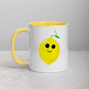 May include: White ceramic mug with a yellow rim and handle. The mug features a cartoon lemon with a smiling face and big eyes.