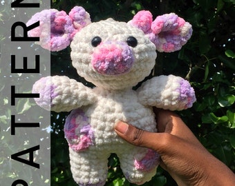 May include: A white crocheted stuffed animal with pink and purple accents. The animal has a rounded body, short legs, and a small, rounded nose. The animal is being held by a hand.