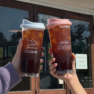 May include: Two clear plastic tumblers with lids, filled with iced coffee. One tumbler has a white lid and says "Dreamer, my head is in the clouds & I'd like to stay here awhile". The other tumbler has a pink lid and says "Coffee Paws" with a cartoon cat and two cartoon bears.