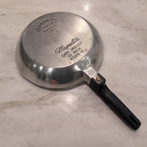 May include: A silver-colored Wagner Ware Magnalite chef skillet with a black handle. The underside features the text Wagner Ware Sidney, Magnalite, Chef Skillet, 10 Inch, and 4509-P. The metal surface shows signs of use with visible scratches.