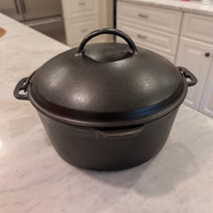 May include: A round black cast iron Dutch oven with a matching lid. The pot features two side handles and a central arched handle on the lid. The surface has a smooth, dark finish, and the design includes a small pouring spout on the side rim.