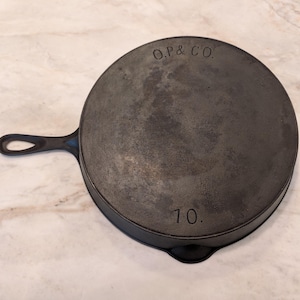 May include: A dark gray cast iron skillet with a single handle. The skillet has the text "O.P. & CO." and "10." embossed on the surface. The pan is round and appears well-used.