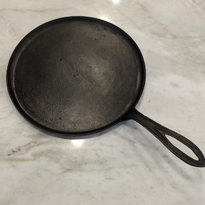 May include: A round, black cast iron griddle pan with a long handle. The pan is circular with a flat cooking surface and a handle with an oval opening. The pan is designed for stovetop cooking.
