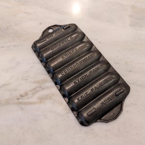 May include: A cast iron Griswold stick pan with six cylindrical molds. The pan is dark gray and has the words "GRISWOLD", "CRISPY", "CORNBREAD", "STICK PAN", and "ERIE PA. U.S.A." embossed on it. The pan is marked "No. 202" and "825".