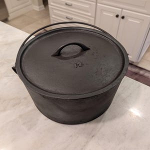 May include: A round black cast iron Dutch oven with a matching lid featuring a central handle and the number 12 embossed on the surface. The pot includes a curved metal bail handle attached to the sides for lifting and carrying the heavy cookware.