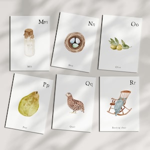 Puede incluir: Ilustraciones en acuarela de tarjetas del alfabeto. Las tarjetas presentan imágenes de una botella de leche, un nido con huevos, aceitunas, una pera, una codorniz y una mecedora. Cada tarjeta tiene una letra y la palabra para la imagen.