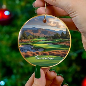 May include: A round, gold-edged Christmas ornament featuring a golf course scene with the word "Temeculah" in green. The ornament is held against a backdrop of a Christmas tree with red ornaments.
